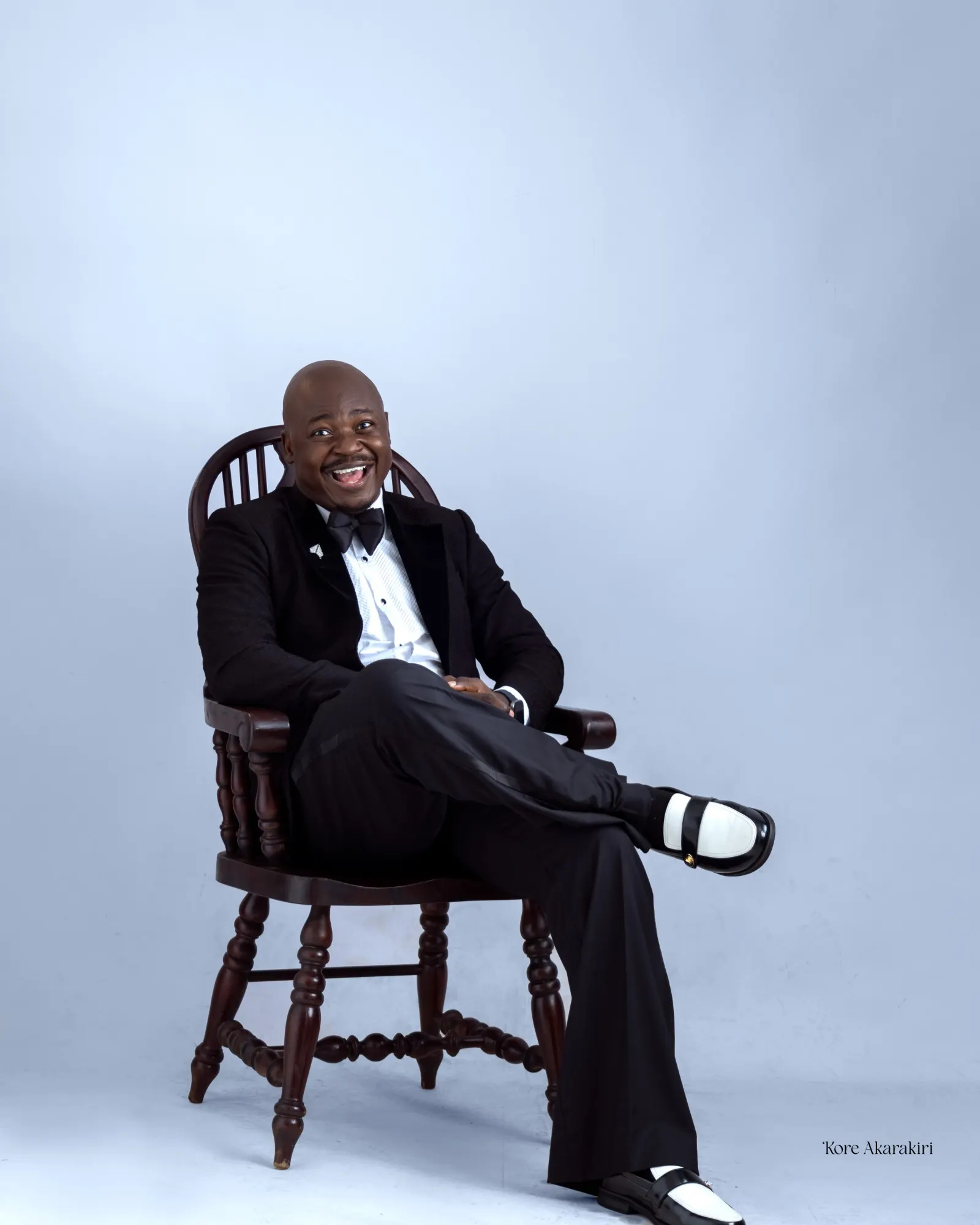 The Corporate Emcee laughing in a tuxedo and bow tie, seated on a wooden chair in a studio portrait