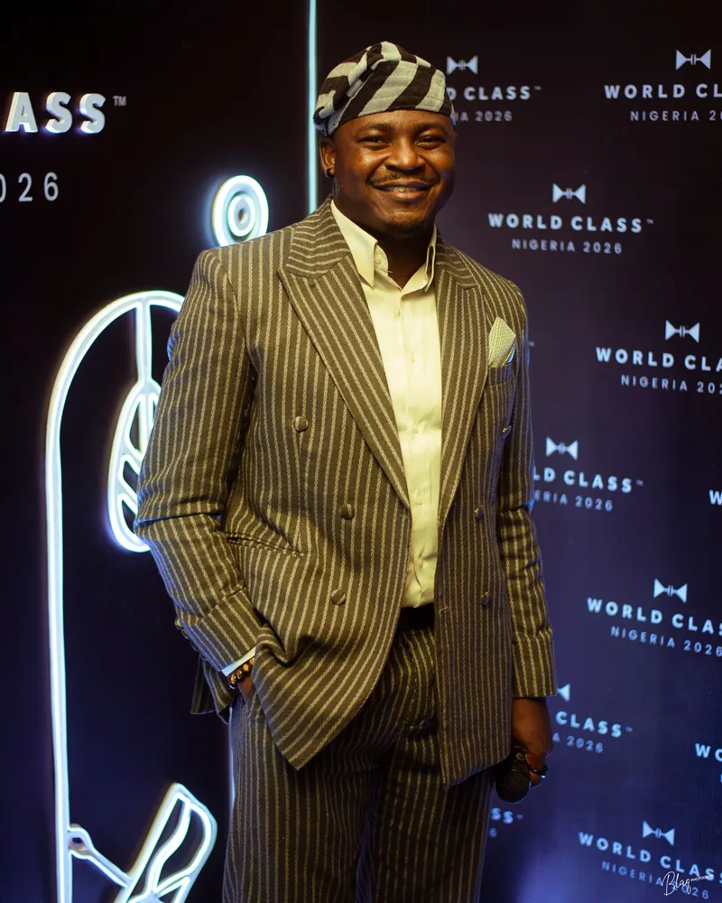 The Corporate Emcee in a pinstripe suit and fila, smiling at the World Class Nigeria 2026 backdrop
