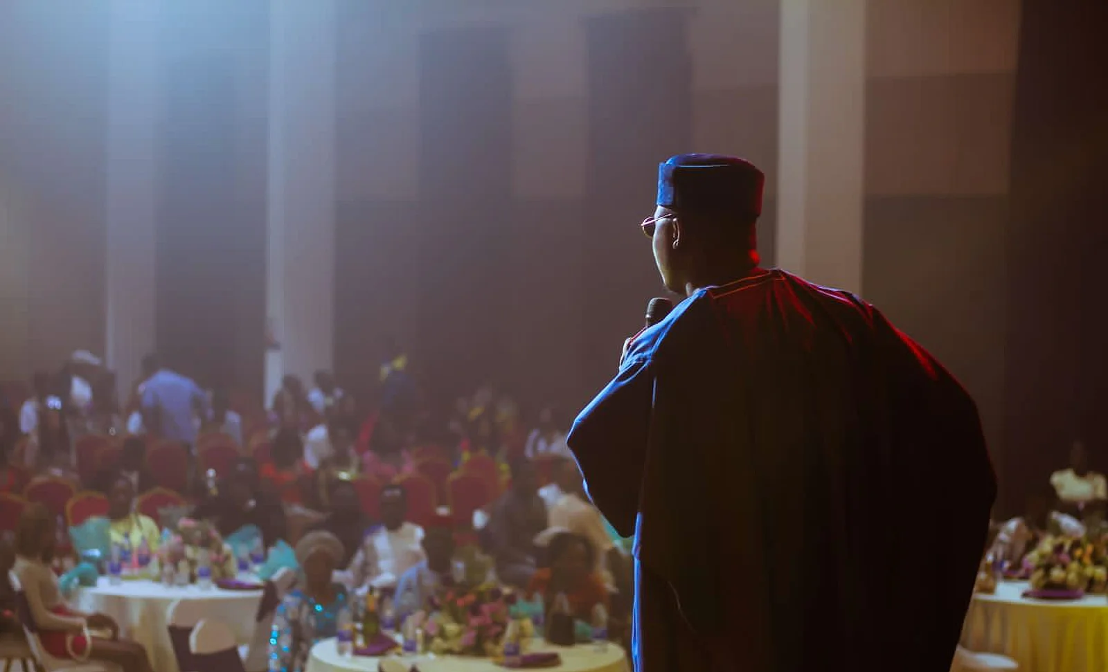 The Corporate Emcee on stage in agbada and fila, microphone in hand, facing a full banquet hall